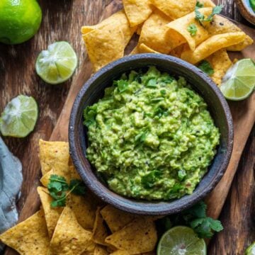 Texas guacamole served with tortilla chips, fresh lime, cilantro, and avocado on a wooden board.