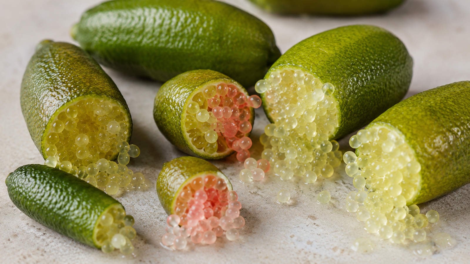 Australian finger limes on a stone surface, with two cut open showing pale green and pink citrus pearls.